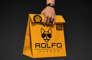 Bolsa de papel naranja de Rolfo Burger con el logotipo del bulldog con gafas, una pegatina de huella y un código QR